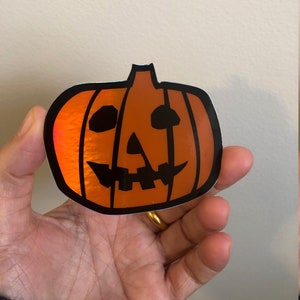 May include: A holographic orange pumpkin sticker with a black outline and a jack-o'-lantern face.