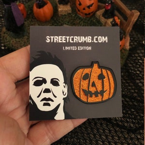 May include: Two enamel pins, one featuring a black and white silhouette of Michael Myers' mask from the Halloween franchise, and the other a glittery orange pumpkin with a black jack-o'-lantern face. The pins are on a black card with the text "STREETCRUMB.COM LIMITED EDITION".