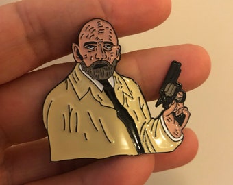 Puede incluir: Un pin de esmalte en blanco y negro de un hombre con barba que lleva un abrigo beige y sostiene un arma. El pin es una ilustración de estilo de dibujos animados.