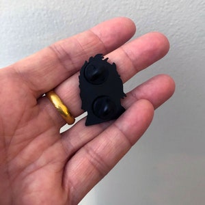 May include: Black enamel pin featuring a silhouette of a person's head with a crown on top. The pin has two metal posts on the back for attaching to clothing or a bag.