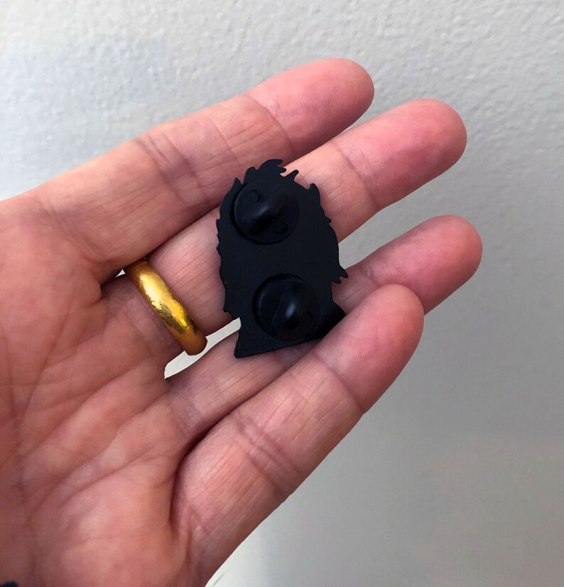 May include: Black enamel pin featuring a silhouette of a person's head with a crown on top. The pin has two metal posts on the back for attaching to clothing or a bag.