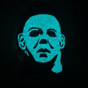 May include: A close-up of a teal blue glow-in-the-dark mask with a human face. The mask has a simple design with a wide mouth and eyes.