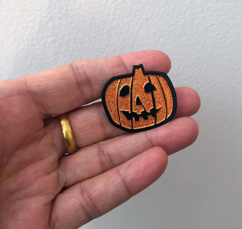May include: A black and orange glitter pumpkin pin with a carved jack-o'-lantern face.