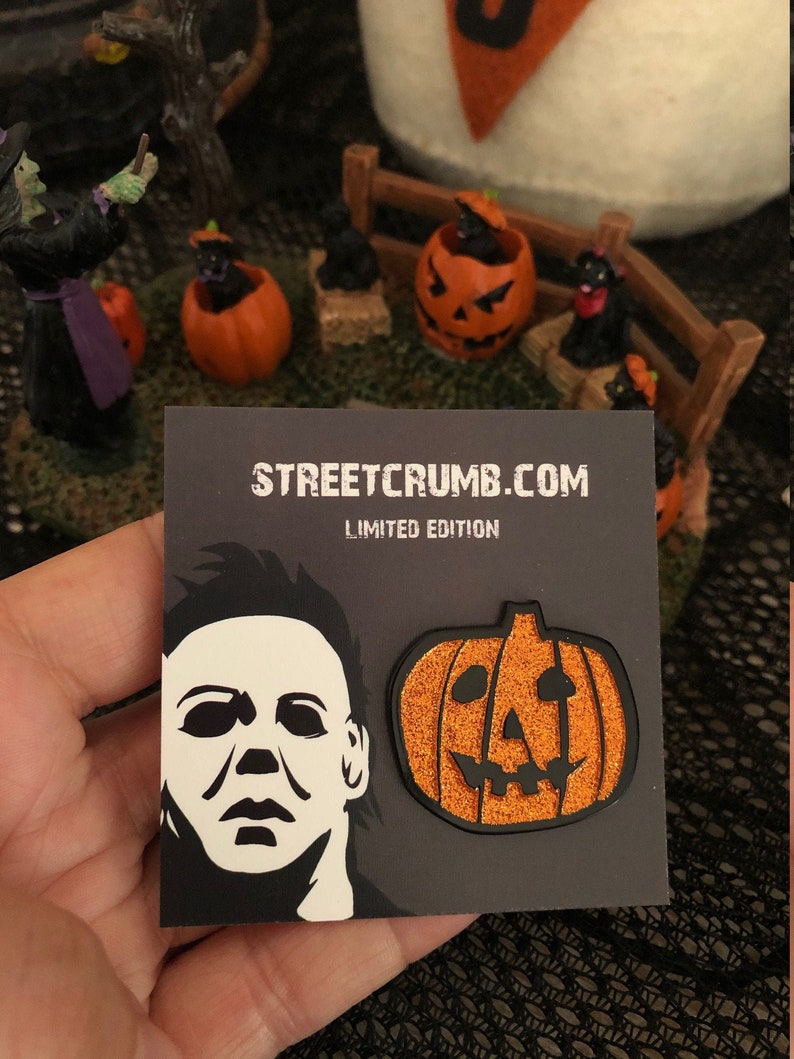 May include: Two enamel pins, one featuring a black and white silhouette of Michael Myers' mask from the Halloween franchise, and the other a glittery orange pumpkin with a black jack-o'-lantern face. The pins are on a black card with the text "STREETCRUMB.COM LIMITED EDITION".