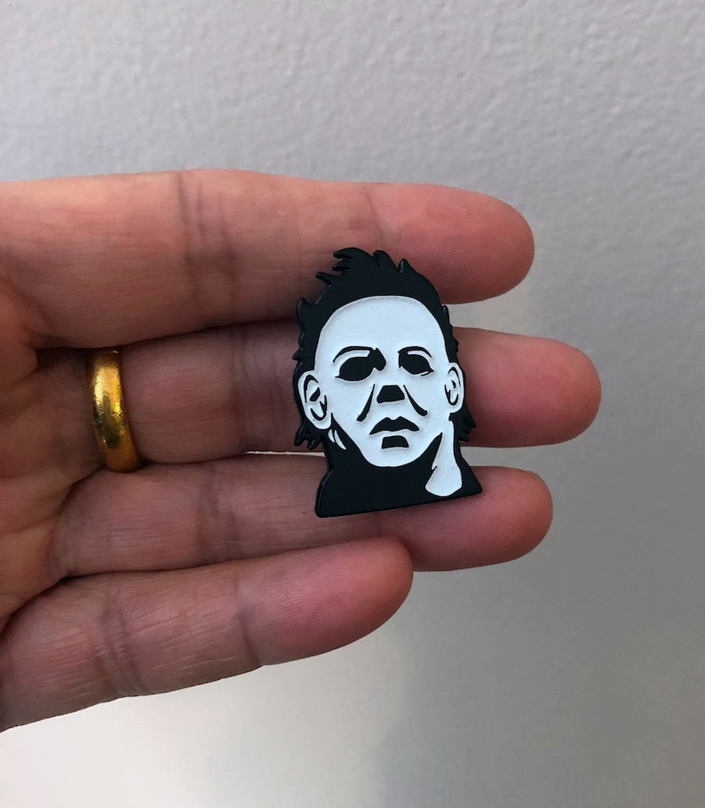 May include: Black and white enamel pin featuring a close-up of Michael Myers' mask from the Halloween franchise. The pin is a popular collectible for horror movie fans.