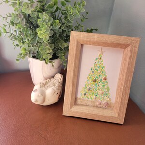 Può includere: Un'opera d'arte incorniciata di un albero di Natale, decorato con conchiglie, in una cornice di legno chiaro. Un piccolo ornamento a forma di uccello bianco e una pianta in vaso sono visibili nell'immagine.