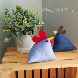 Puede incluir: Dos almohadas de pollo de fieltro, una azul con una cresta y un pico marrones, la otra azul claro con una cresta roja y un pico amarillo. Las almohadas están sentadas en una caja de madera. El texto "Whimsy Wish Designs" es visible en el fondo.