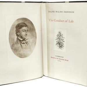 Puede incluir: Libro abierto con cubierta negra. La p&aacute;gina izquierda presenta un retrato en sepia de Ralph Waldo Emerson en un marco ovalado. La p&aacute;gina derecha muestra el t&iacute;tulo "The Conduct of Life" y una peque&ntilde;a ilustraci&oacute;n de un &aacute;rbol. Impreso en Cambridge en 1904.