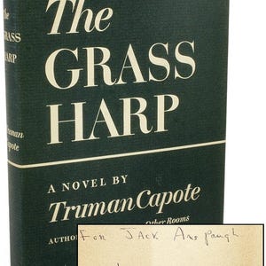 Könnte beinhalten: Ein grünes Hardcover-Buch mit dem Titel "The Grass Harp" von Truman Capote. Das Buch hat eine handschriftliche Widmung auf dem vorderen Umschlag, die lautet "For Jack Ans Paugh from Truman Capote".