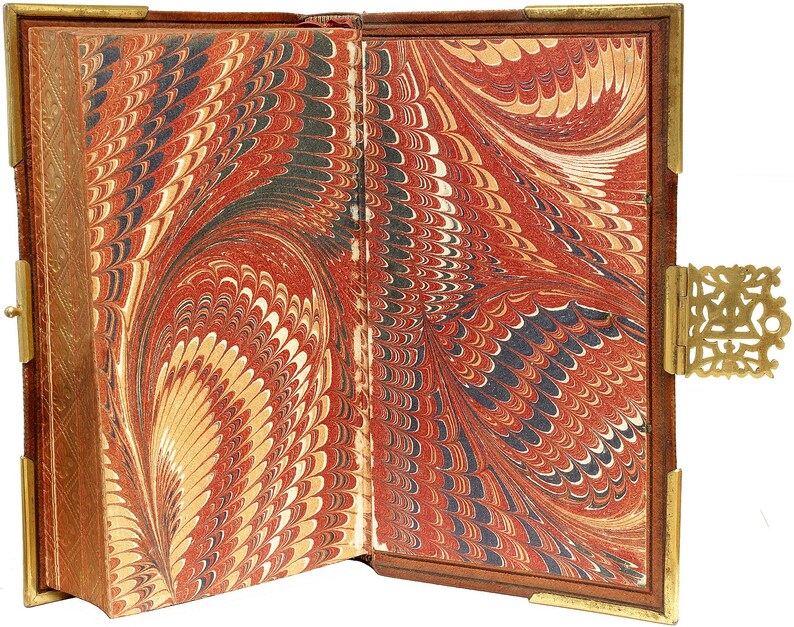 Puede incluir: Libro antiguo abierto con p&aacute;ginas de papel jaspeado en tonos rojo, negro y crema. El libro tiene una cubierta de cuero marr&oacute;n con protectores de esquina de metal dorado y un cierre decorativo. Las p&aacute;ginas muestran un patr&oacute;n arremolinado.