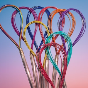 May include: A collection of colorful wire whisks with looped handles. The whisks feature a variety of vibrant colors, including red, orange, yellow, green, blue, and purple. The handles are made of metal and are attached to the wire loops.