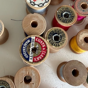 May include: A collection of vintage wooden spools of thread in various colors, including red, yellow, pink, and blue. Some spools have labels with text like "J&P Coats" and "Heavy Duty". The spools are arranged on a light-colored surface.