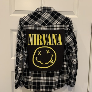 May include: A long-sleeve button-down shirt featuring a black and white plaid pattern. The back of the shirt displays a large rectangular black patch with the yellow text NIRVANA printed above a yellow graphic of a smiley face with x-shaped eyes.