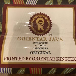 May include: A rectangular fabric label with a white background and dark red border. The text reads ORIENTAR JAVA, 100% COTTON, 6 YARDS, 5.486 METRES, ORIGINAL, PRINTED BY ORIENTAR KINGTEX. A circular logo with the letters OR is centered at the top.