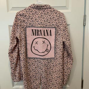 May include: A long-sleeved button-down shirt in a light pink fabric with a small dark blue floral pattern. The back features a large, square, light pink patch with a scalloped gray border, displaying the black text NIRVANA and a smiley face.
