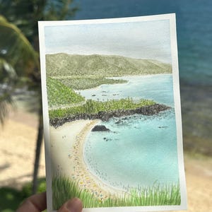 May include: A watercolor painting of a coastal scene featuring a sandy beach, turquoise water, and green vegetation. The artwork is held up in front of a window overlooking a similar ocean view, with a blue sky and palm trees visible.