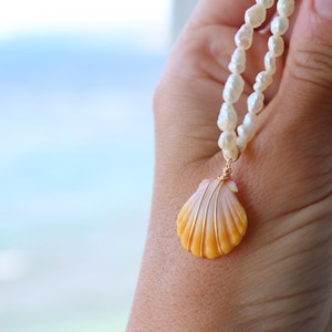 Può includere: Una collana fatta a mano con un ciondolo a forma di conchiglia. La conchiglia, nei toni dell'arancione e del giallo, è avvolta in filo d'oro e appesa a una catena di perline bianche di forma irregolare. Lo sfondo mostra una vista sfocata del mare.