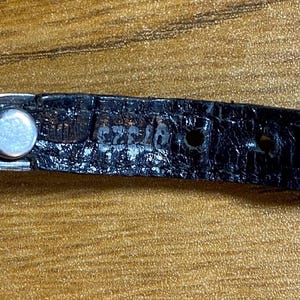 May include: Close-up of a black leather watch strap with a silver buckle. The strap has a textured surface and the buckle has a small, round, silver button.