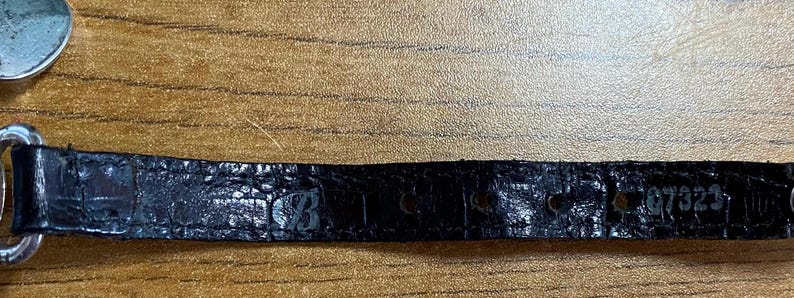 May include: Black leather watch band with a crocodile pattern and the number "87323" embossed on it.