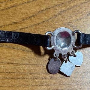 May include: A black leather bracelet with a silver charm featuring a round, clear glass stone. The charm has three smaller silver charms attached to it.