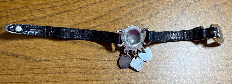 May include: A black leather bracelet with a silver charm featuring a round, clear glass stone. The charm has three smaller silver charms attached to it.