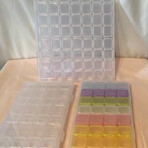 May include: Three clear plastic storage boxes with compartments. The top box has 48 compartments, the middle box has 36 compartments, and the bottom box has 24 compartments. The bottom box has compartments in four rows of six, with each row a different color: purple, green, pink, and yellow.