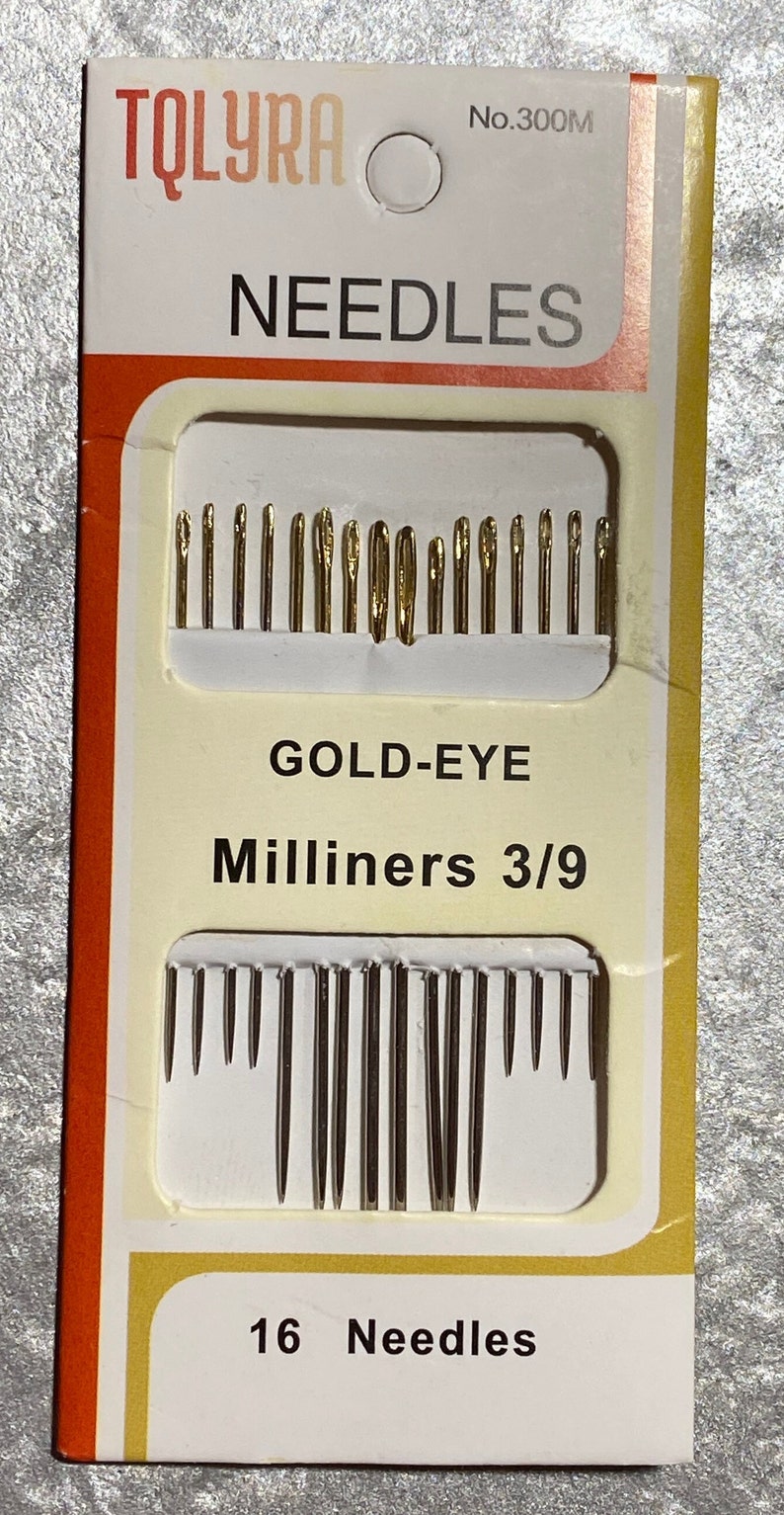 May include: A package of gold-eye milliners needles. The package is white with orange and yellow accents. The text on the package reads "TQLYRA", "NEEDLES", "GOLD-EYE Milliners 3/9", and "16 Needles". The needles are arranged in two rows.