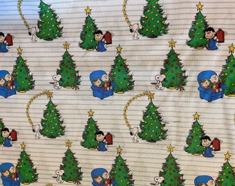 May include: A white fabric with a repeating pattern of Christmas trees, Snoopy, and Charlie Brown. The characters are dressed in red and blue holiday attire. The background is a light blue with white stripes.