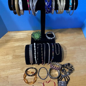 May include: A collection of bracelets in various styles and colors displayed on a black jewelry stand. The bracelets include bangles, beaded, and chain styles, with accents of silver, gold, blue, and red. Some bracelets have decorative elements like gems and enamel.