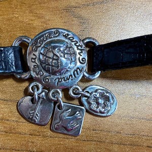May include: A black leather bracelet with a silver charm that says "Love our Earth" and features a globe. The bracelet also has several other silver charms, including a heart with an arrow, a dove, and a flower.