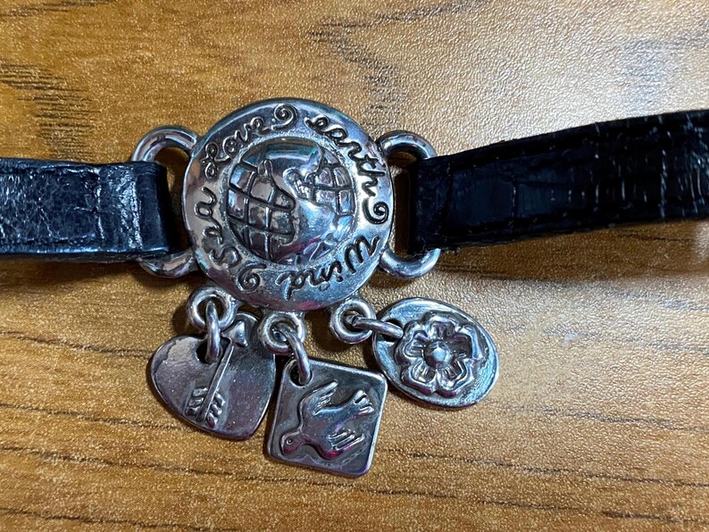 May include: A black leather bracelet with a silver charm that says "Love our Earth" and features a globe. The bracelet also has several other silver charms, including a heart with an arrow, a dove, and a flower.