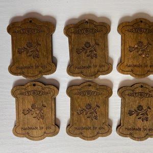 May include: Six wooden tags with a floral design and the text "Handmade for you" are arranged on a white surface. The tags are a warm brown color and have a decorative border.