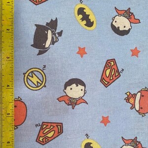May include: Light blue fabric featuring cartoon illustrations of Batman, Superman, and The Flash, along with their logos and orange stars. The fabric is measured with a yellow ruler showing inches.