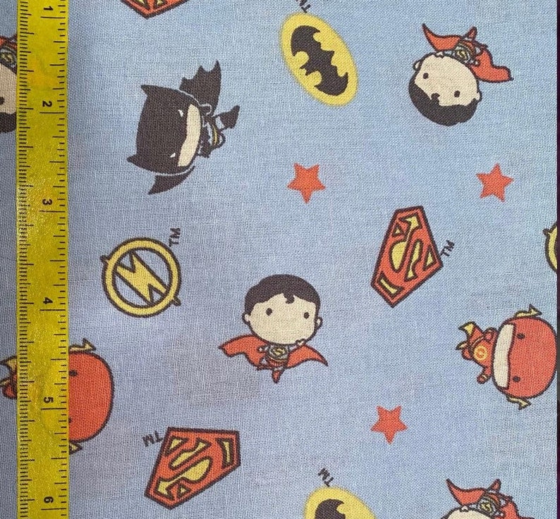 May include: Light blue fabric featuring cartoon illustrations of Batman, Superman, and The Flash, along with their logos and orange stars. The fabric is measured with a yellow ruler showing inches.