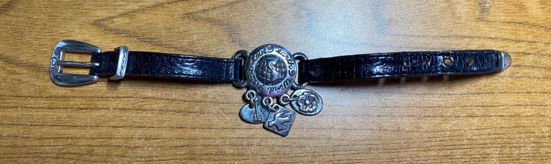May include: A black leather bracelet with a silver charm that says "Loved by earth" and has a dove design. The bracelet has a silver buckle.