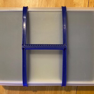 Może przedstawiać: Prostokątny, przezroczysty plastikowy pojemnik z niebieskim uchwytem i niebieskim mechanizmem zatrzaskowym. Pojemnik przeznaczony jest do przechowywania i organizacji. Pokrywa jest półprzezroczysta, co pozwala na widoczność zawartości.