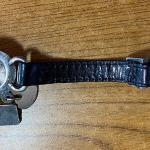 May include: A silver charm bracelet with a black leather strap and a variety of silver charms, including a round charm with a green and blue design.