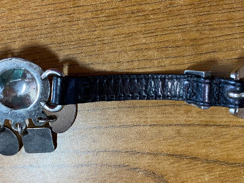 May include: A silver charm bracelet with a black leather strap and a variety of silver charms, including a round charm with a green and blue design.