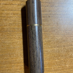 May include: A cylindrical wooden container with a dark brown finish and a gold-colored band. The container is made of wood and has a smooth, polished surface. The design is simple and elegant.