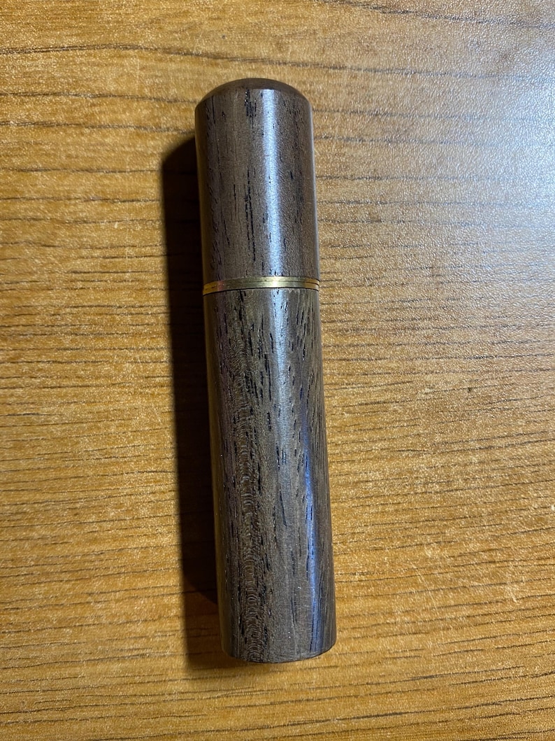 May include: A cylindrical wooden container with a dark brown finish and a gold-colored band. The container is made of wood and has a smooth, polished surface. The design is simple and elegant.