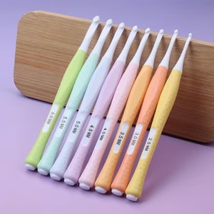 May include: A set of eight crochet hooks in various pastel colors, including green, blue, pink, orange, and yellow. Each hook has a white metal tip and a labeled handle with sizes ranging from 2.5 mm to 6.0 mm. They are arranged on a wooden surface.