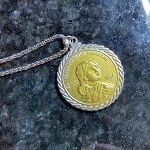 Op de afbeelding: Een goudkleurige hangerketting met een munt met een portret van een inheemse Amerikaanse vrouw. De munt is gegraveerd met "REMEMBER NATIVE AMERICANS" en "2016". De ketting heeft een zilveren ketting.
