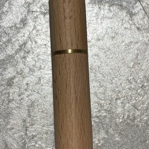 May include: A cylindrical wooden needle case with a brass band. The case is made of light-colored wood and has a smooth, polished finish. The brass band adds a touch of elegance to the design.