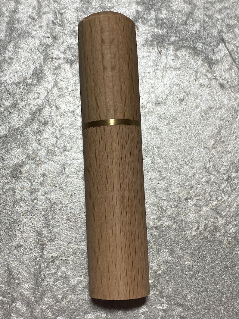 May include: A cylindrical wooden needle case with a brass band. The case is made of light-colored wood and has a smooth, polished finish. The brass band adds a touch of elegance to the design.