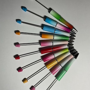 May include: A collection of colorful pens with a gradient design. Each pen has a different color scheme, transitioning from one hue to another. The pens are arranged in a fan shape, showcasing their variety of colors and designs.