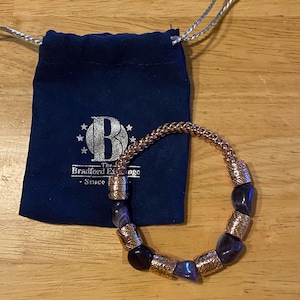 Op de afbeelding: Een koperkleurige armband met donkerblauwe en paarse stenen. De armband ligt naast een donkerblauwe trekkoordtas met de tekst "The Bradford Exchange - Since" erop. De armband ligt op een houten oppervlak.