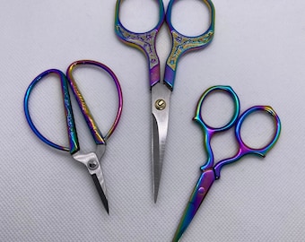 May include: Three pairs of rainbow-colored scissors. The scissors have a metallic finish with hues of purple, blue, green, and gold. One pair has a curved handle, another has a floral design, and the third has a classic design. These are ideal for crafting and sewing.