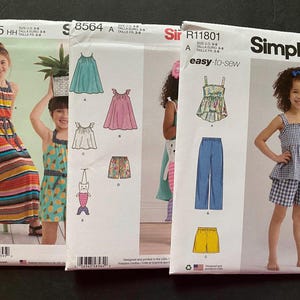 Puede incluir: Tres sobres de patrones de costura Simplicity con diseños de ropa infantil. Los patrones incluyen vestidos, tops, pantalones cortos y pantalones. Los sobres muestran imágenes de las prendas terminadas y el logotipo de Simplicity.