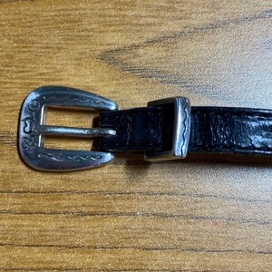 May include: A black leather belt with a silver buckle. The buckle has a floral design and a wavy edge.