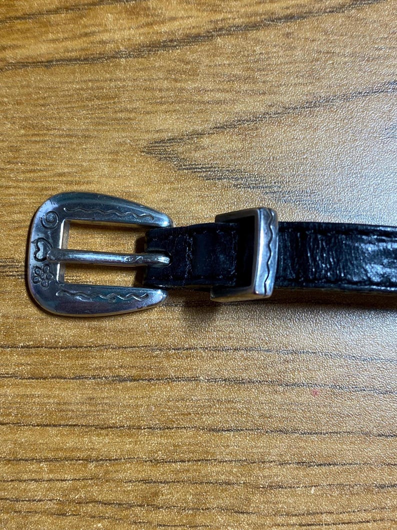 May include: A black leather belt with a silver buckle. The buckle has a floral design and a wavy edge.